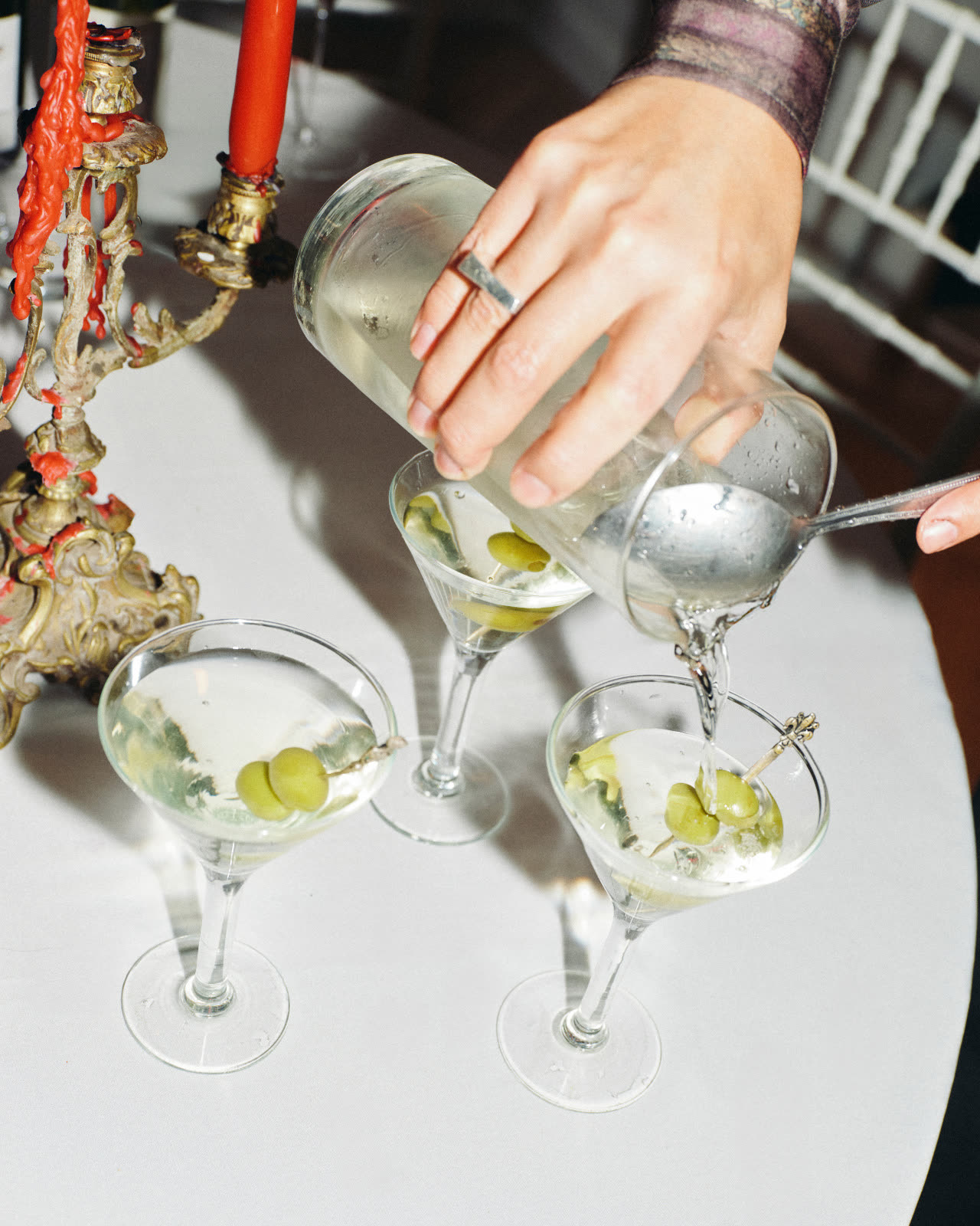 A hand pouring martinis into three coupes on a white cloth, beside a coral candelabra.